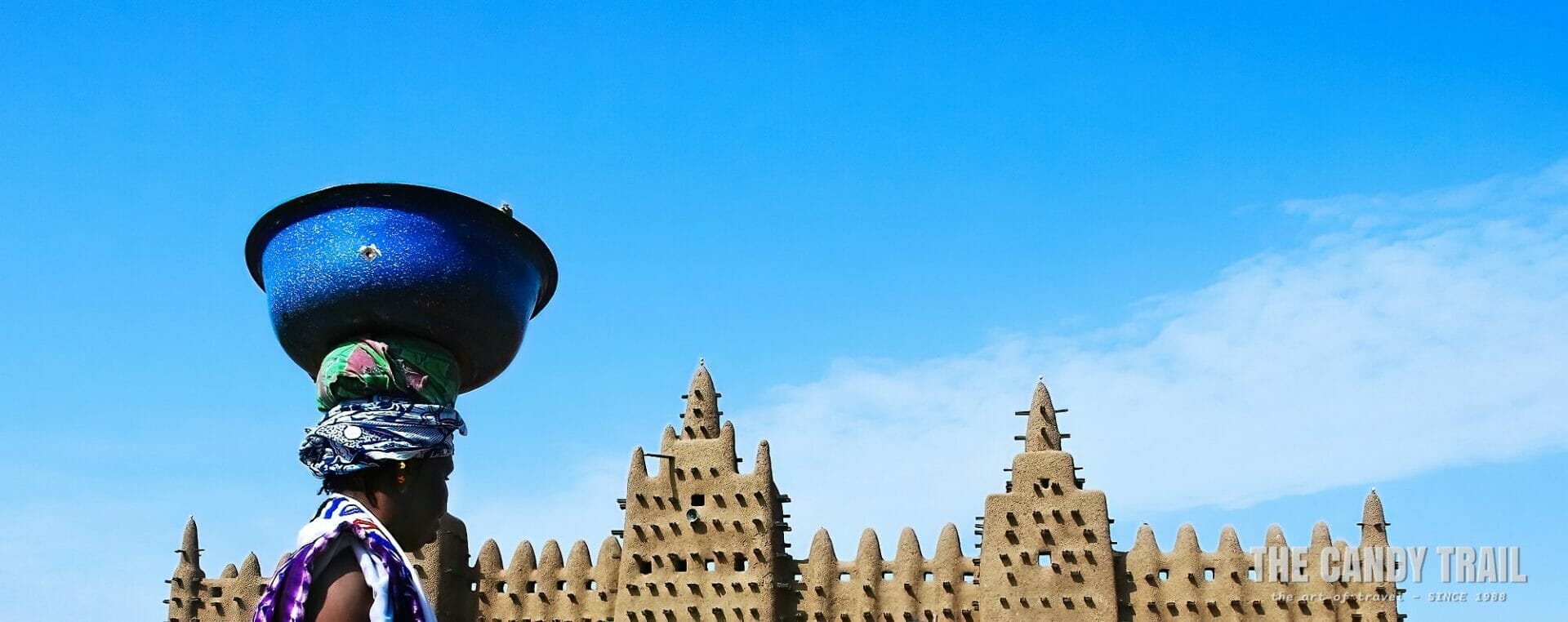 woman with bowl balanced on head walks pass great mosque of djenne in mali.